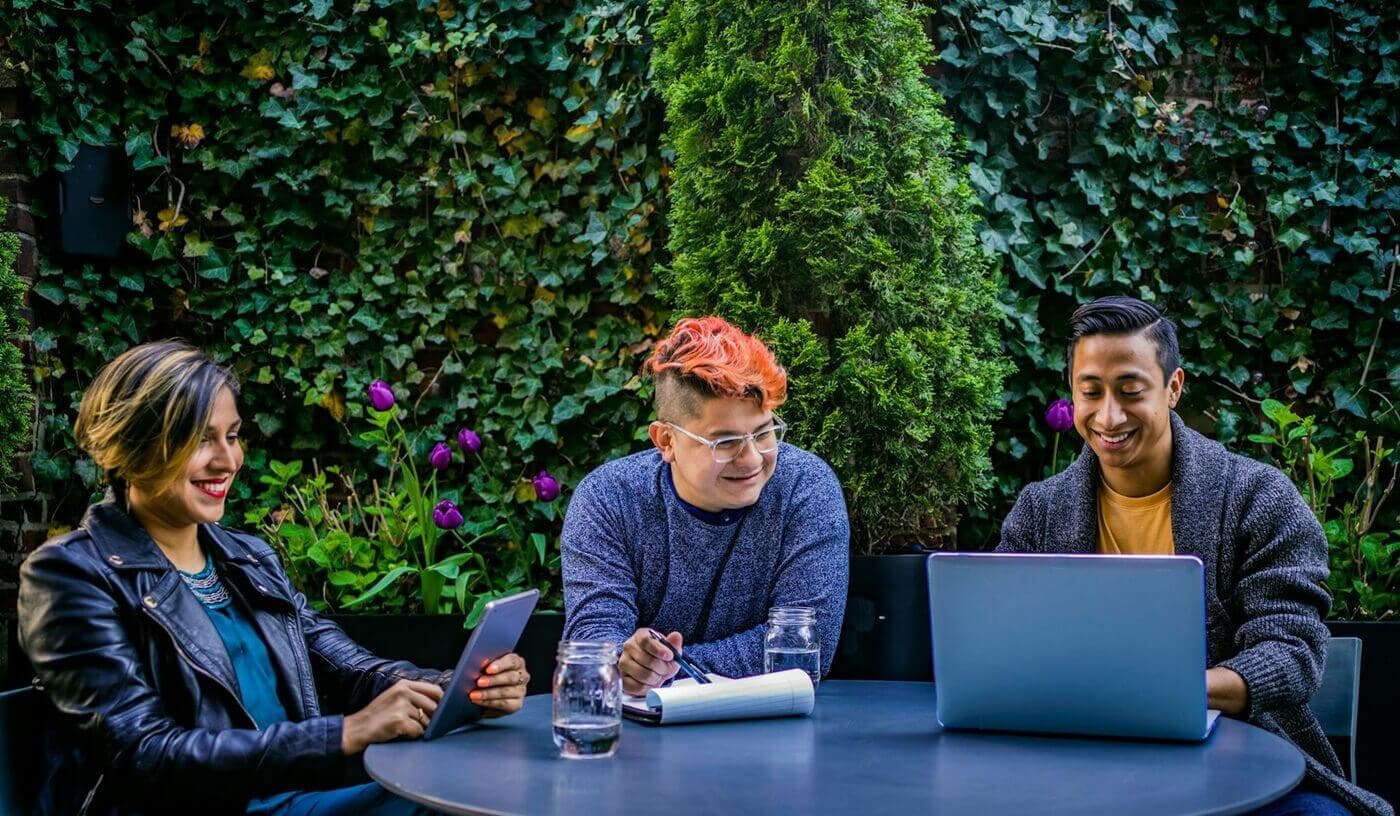 A group of three coworkers sit at an outdoor table discussing work over laptops.