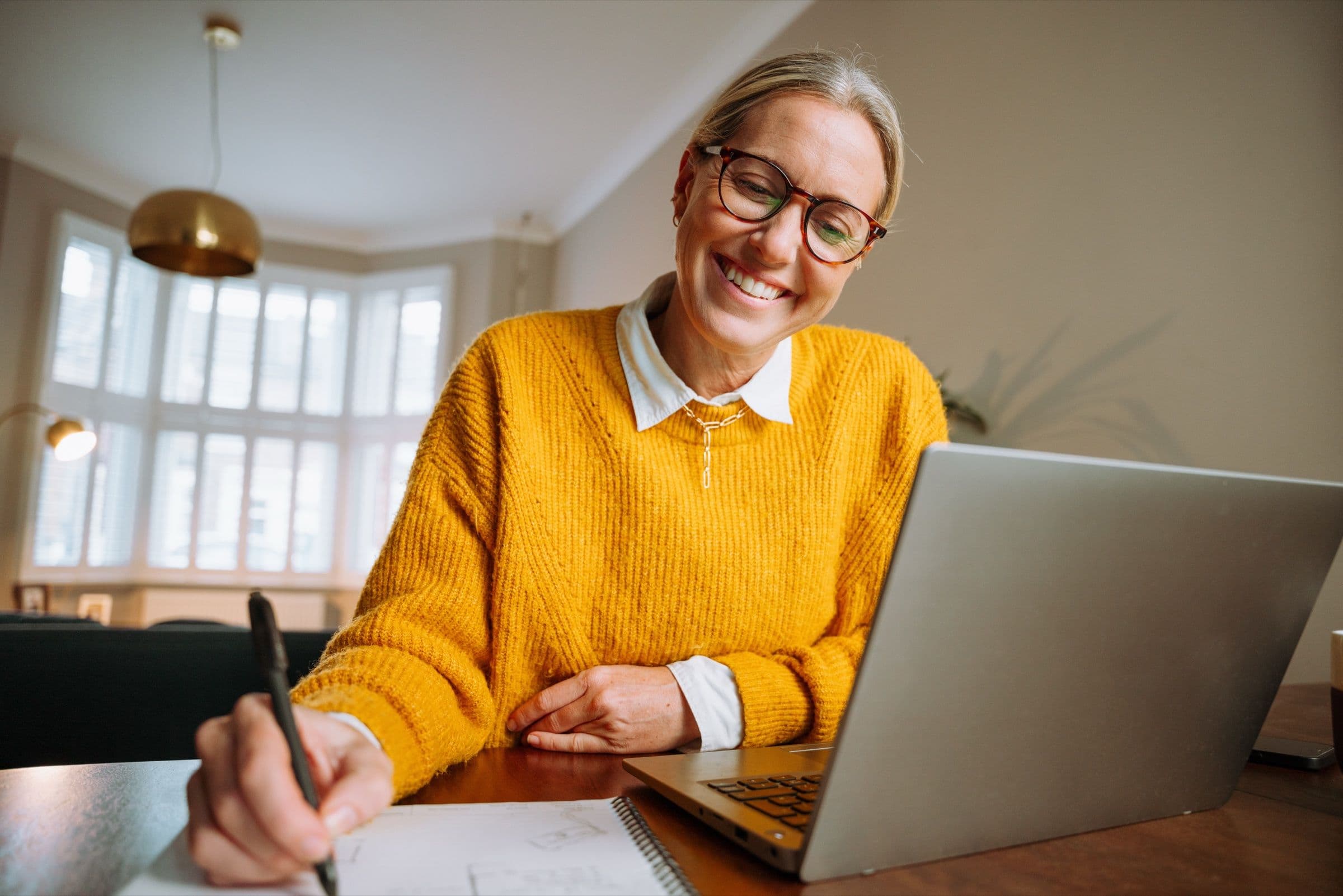A smiling woman in glasses and a yellow sweater takes notes next to her laptop
