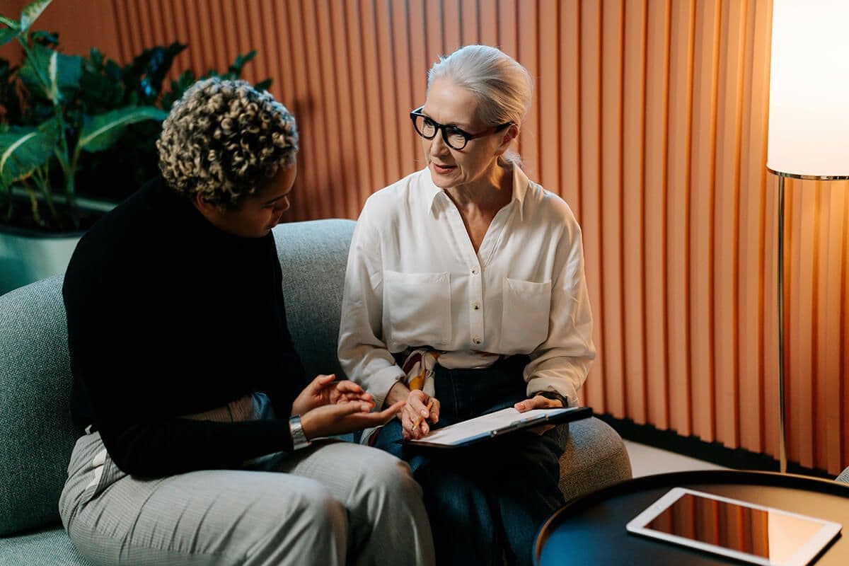 Two women sitting on a couch in a modern office looking at a clipboard. They're discussing the AI tools HR uses.